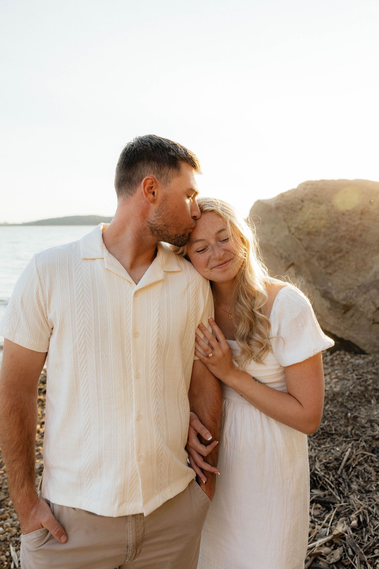 Romantic couple in white outfits embrace on a sunny beach, man kissing woman's forehead near large rock.