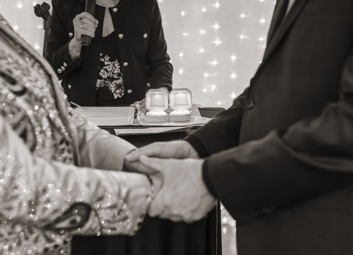 Black and white wedding ceremony photo showing couple holding hands while officiant reads vows with rings displayed nearby.