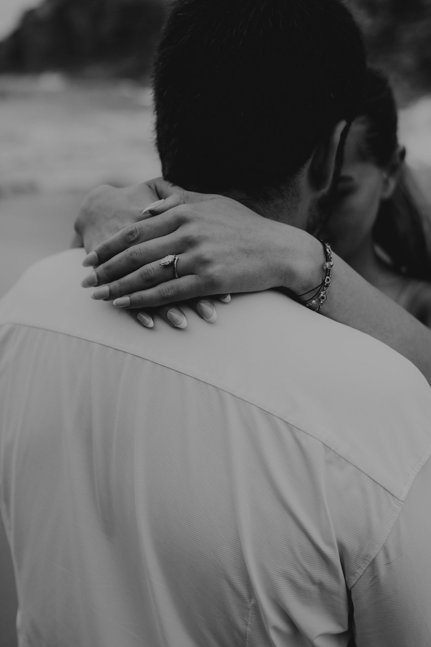 Black and white photo of a couple embracing, woman's hand with ring and bracelet resting on man's shoulder.