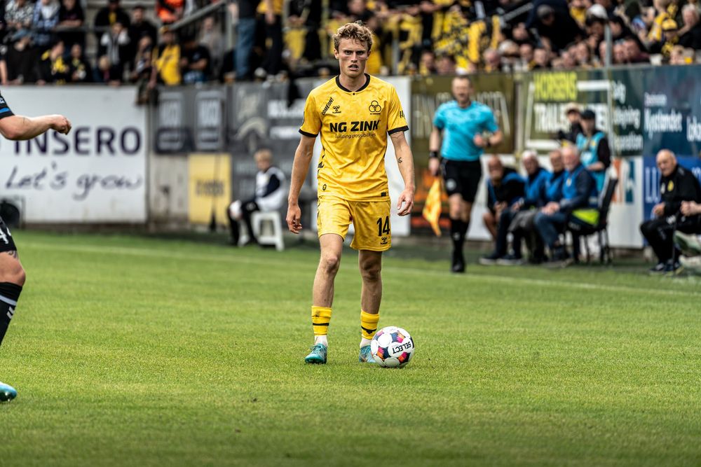 A young footballer in yellow kit dribbles the ball during a competitive match at a packed stadium.