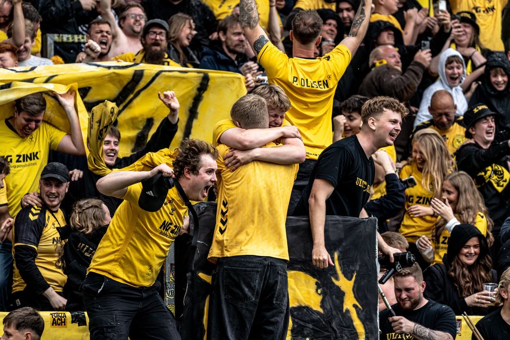 Excited AC Horsens fans in yellow jerseys celebrating together in crowded stadium stands.