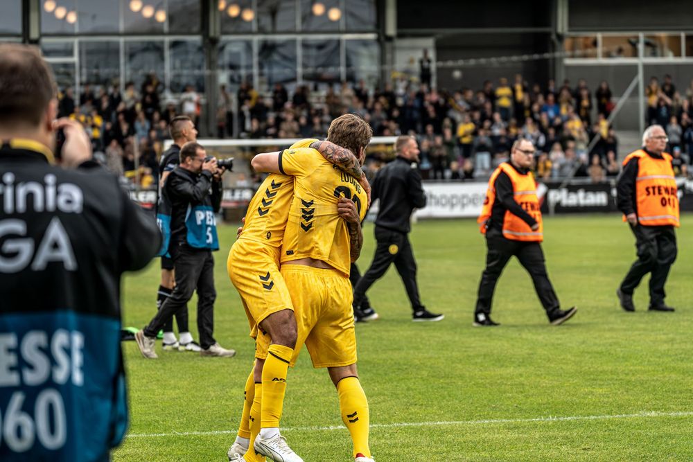 Two soccer players in yellow uniforms embrace on the field celebrating a goal during a match.