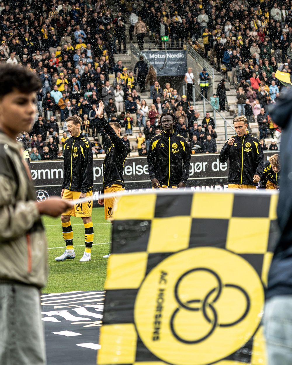 AC Horsens football players line up on the pitch during pre-match ceremony with fans and checkered club flag in foreground.