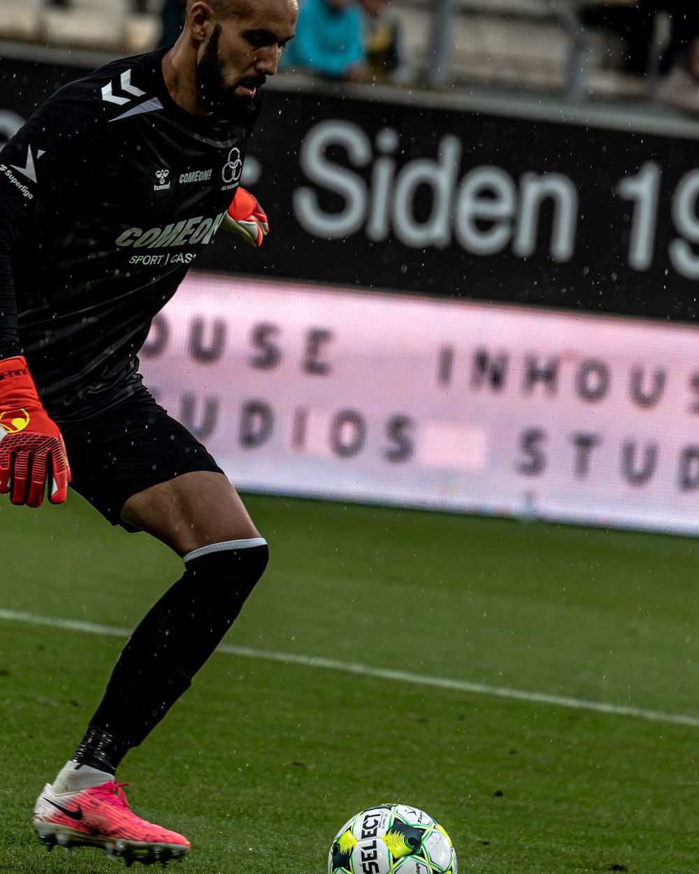 A goalkeeper in black uniform kicks a soccer ball on the field during a match at a stadium.