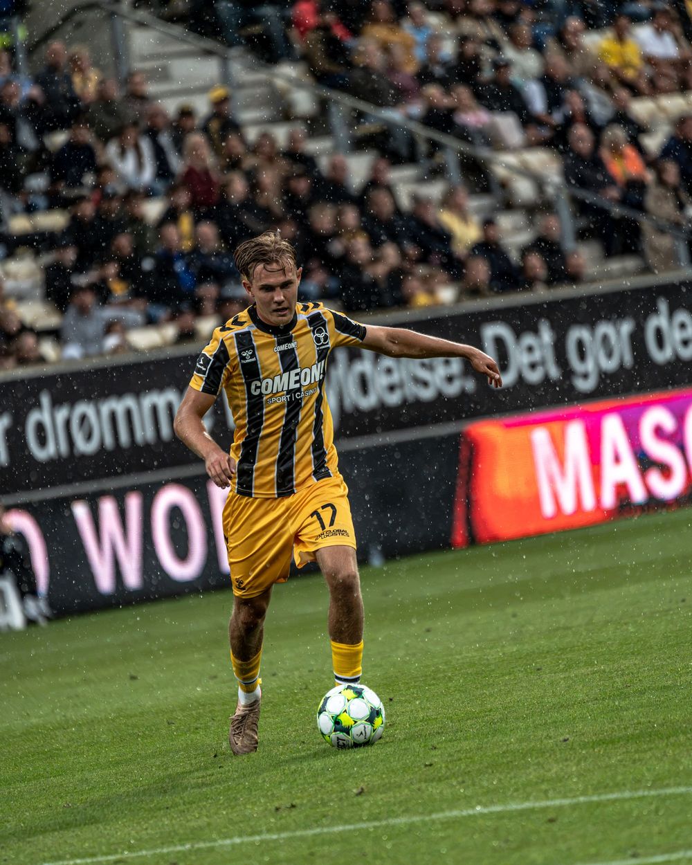 Soccer player in yellow and black striped jersey dribbling ball on wet field during match.
