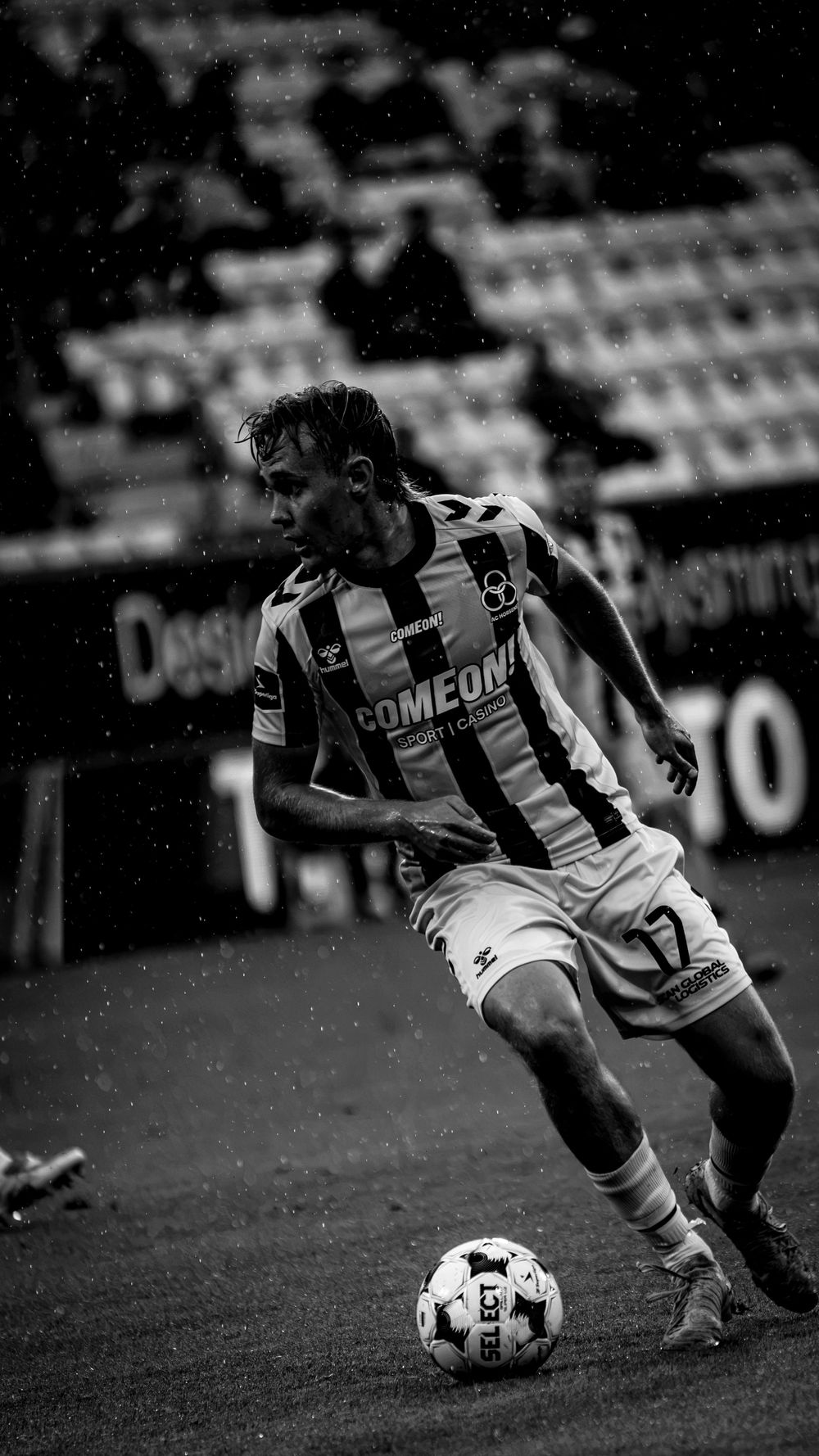 Black and white photo of soccer player in striped kit controlling ball in rainy match.