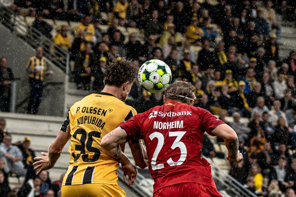 Two soccer players heading the ball during a match, with player 23 Nokheim in red and player 35 in yellow jerseys.