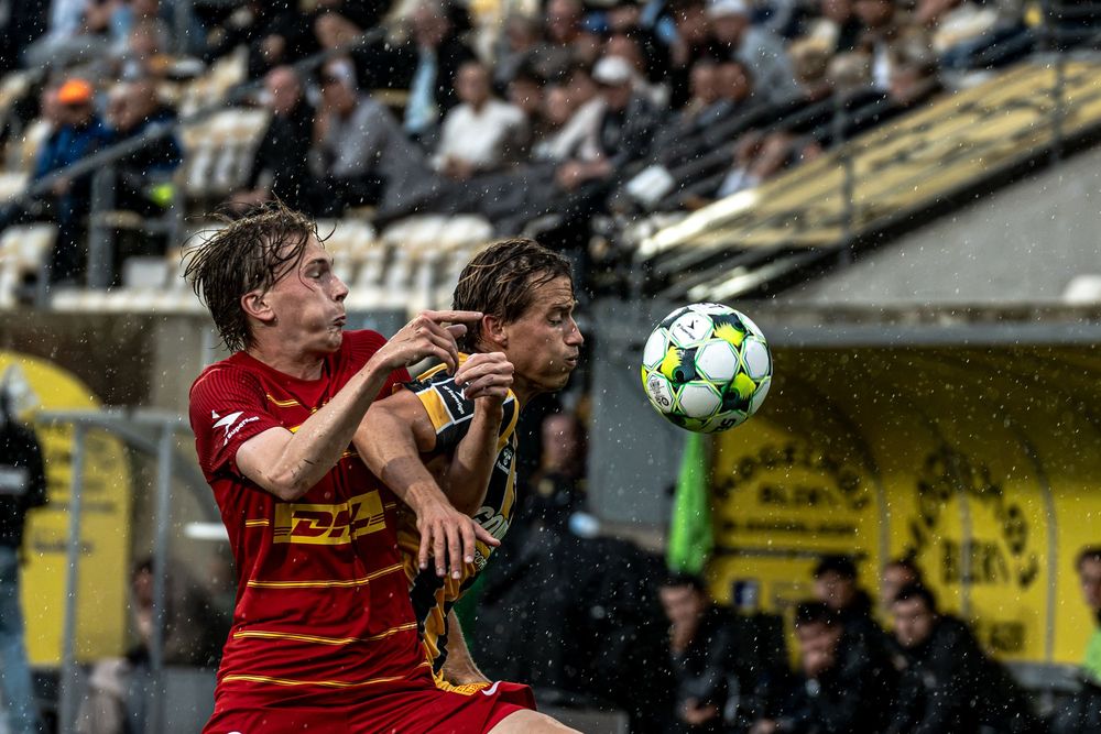 Two soccer players battle for a header during a rainy match, one in red and one in green and yellow jerseys.