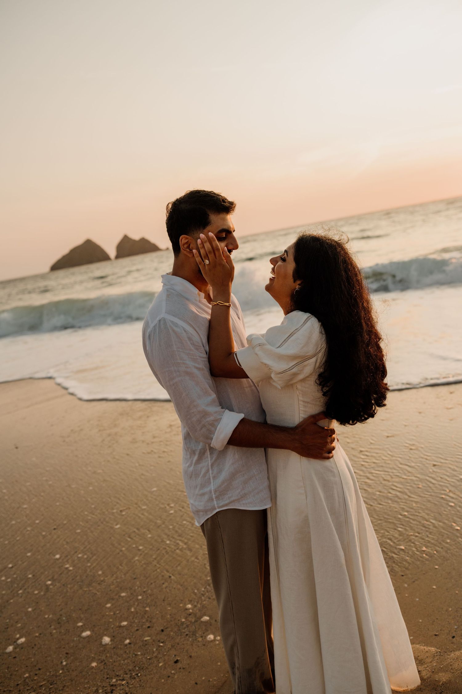 Couple in white outfits embracing romantically on a sandy beach at sunset with ocean waves in the background.