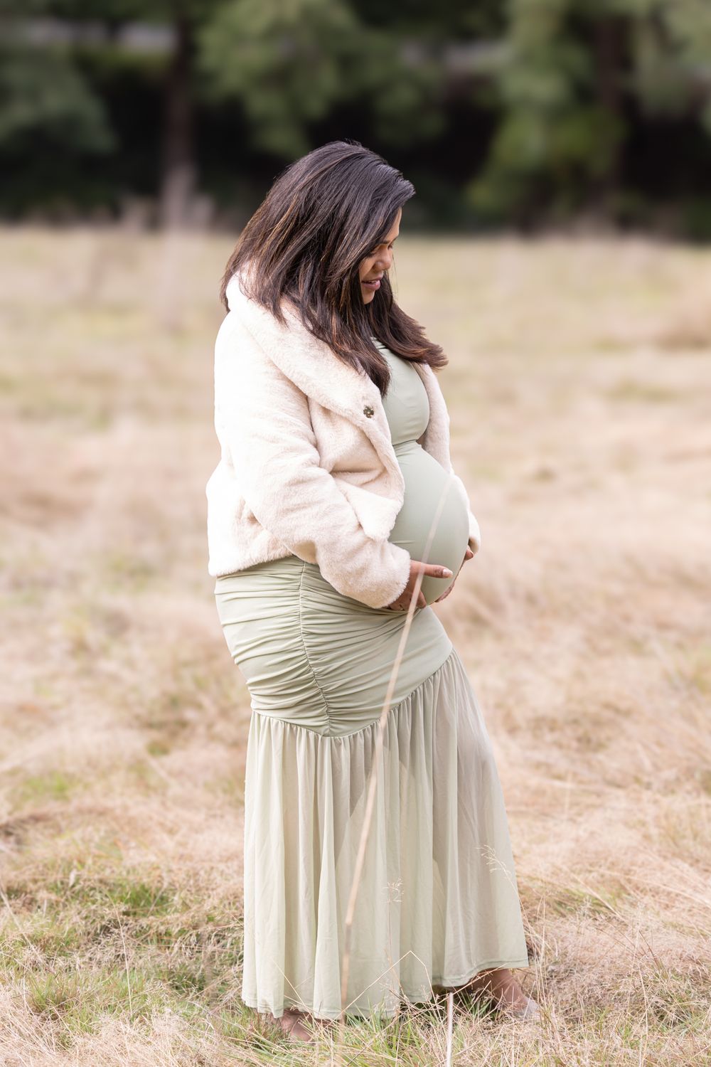 Pregnant woman in sage green dress and cream jacket cradling her baby bump outdoors in a golden field.
