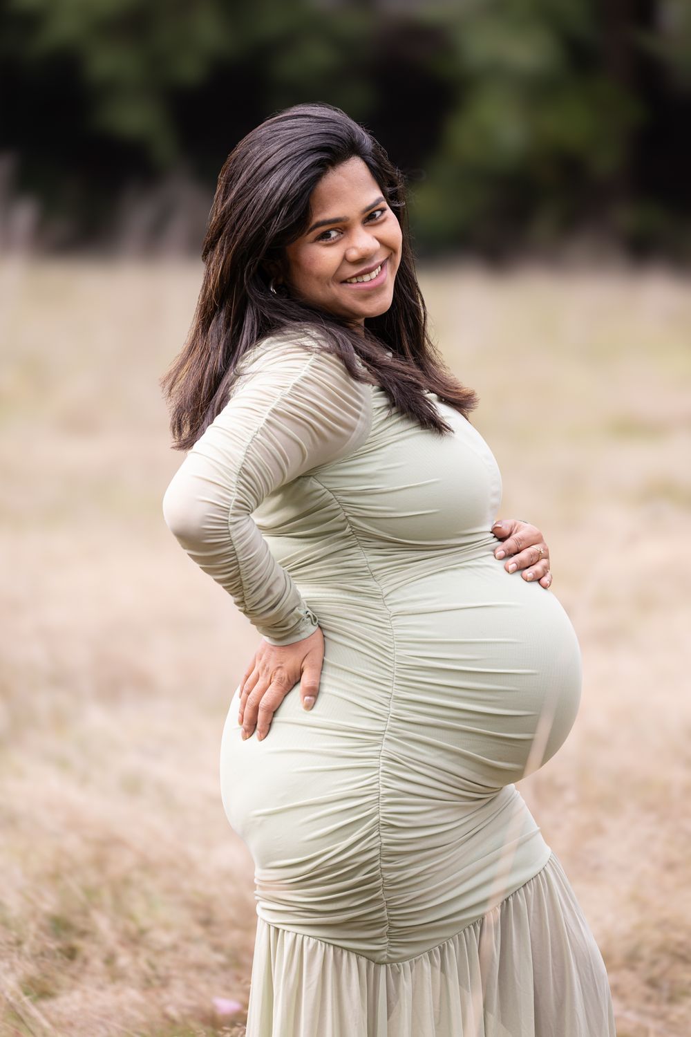 Smiling pregnant woman in sage green ruched dress cradling baby bump outdoors in a field.
