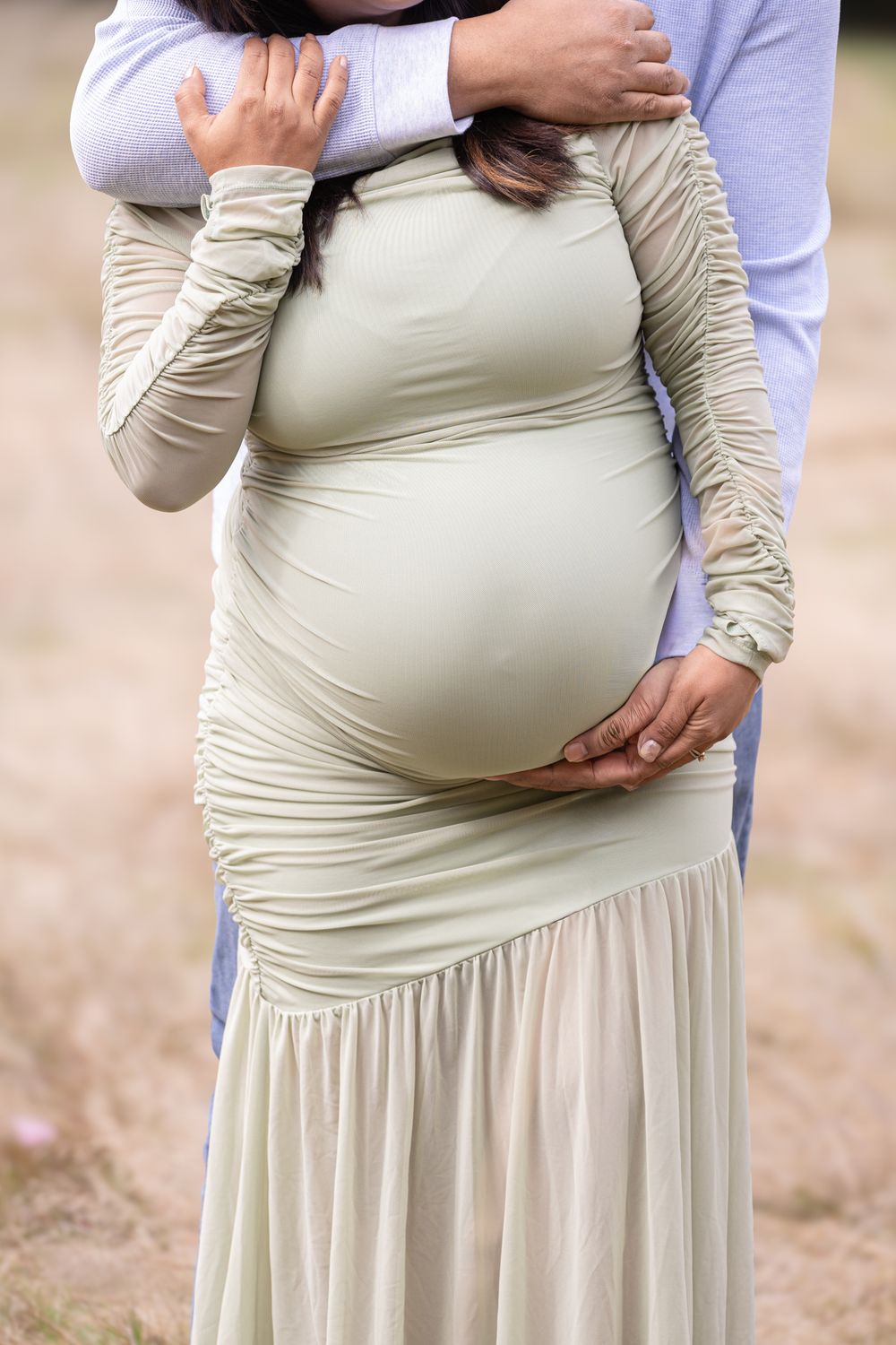 Pregnant woman in sage green ruched dress being embraced by partner outdoors, hands cradling baby bump.