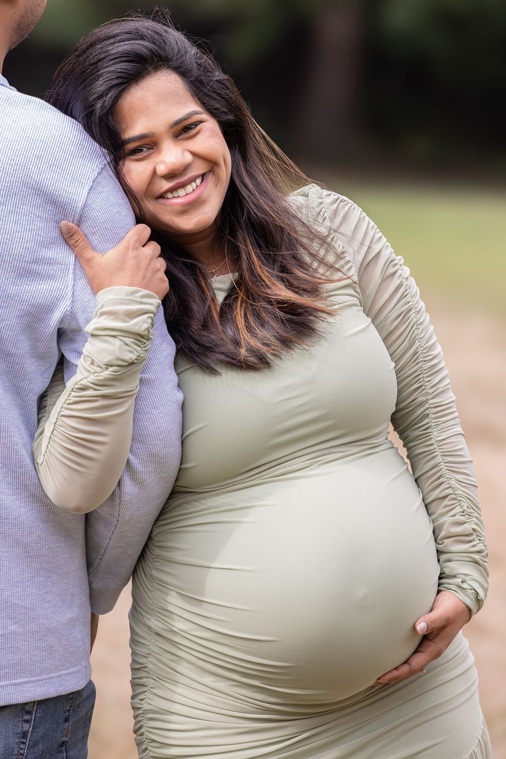Smiling pregnant woman in sage green dress tenderly embraces partner outdoors in a park setting.