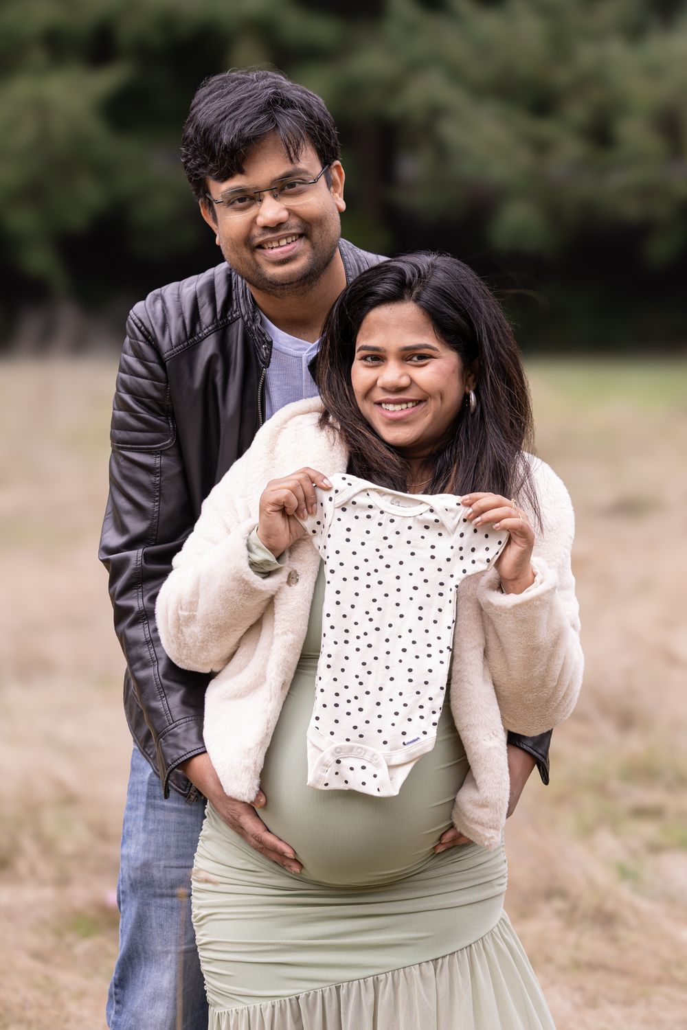 Happy couple outdoors holding baby onesie over pregnant belly, man in leather jacket stands behind woman.