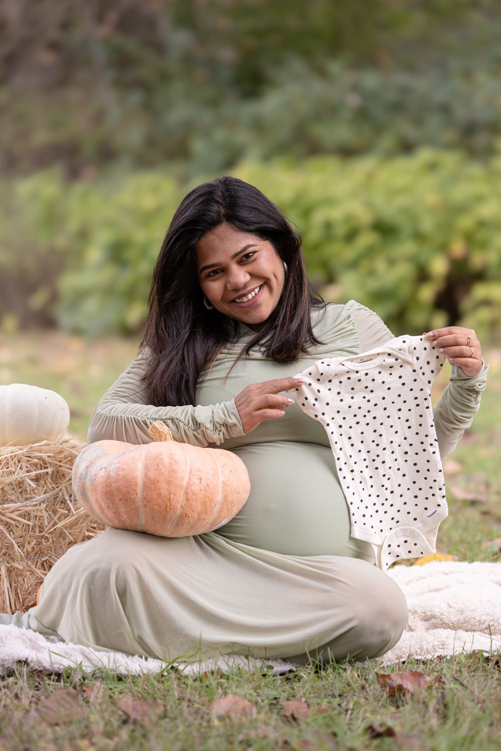Smiling pregnant woman sitting outdoors holding polka dot baby onesie beside pumpkins during fall maternity photoshoot.