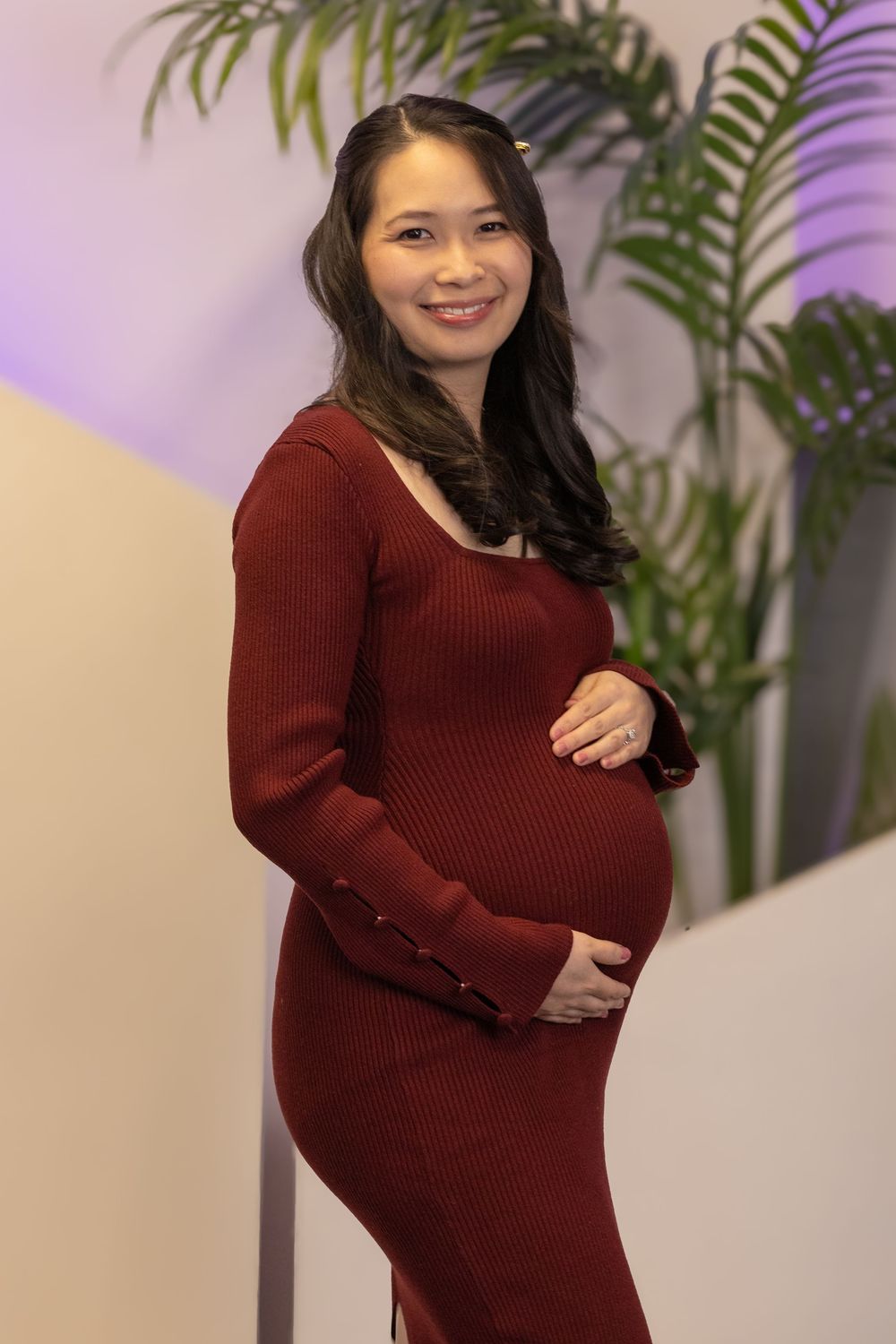 Smiling pregnant woman in red ribbed dress cradling baby bump, posing indoors near tropical plant.