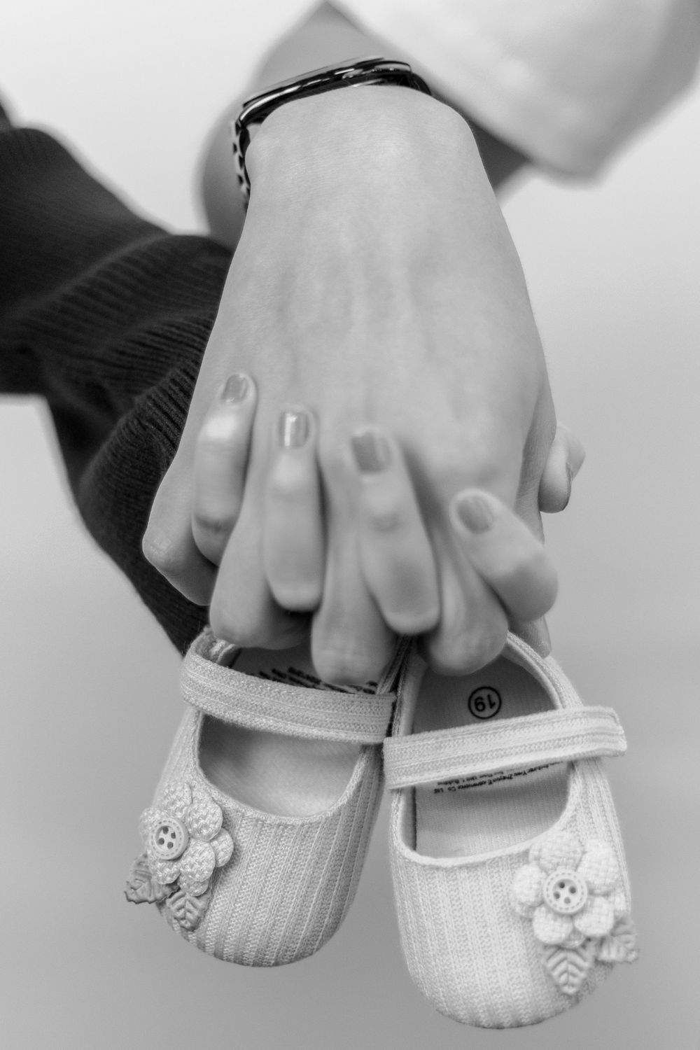 Black and white photo of two adults holding hands over tiny baby girl shoes with flower details.