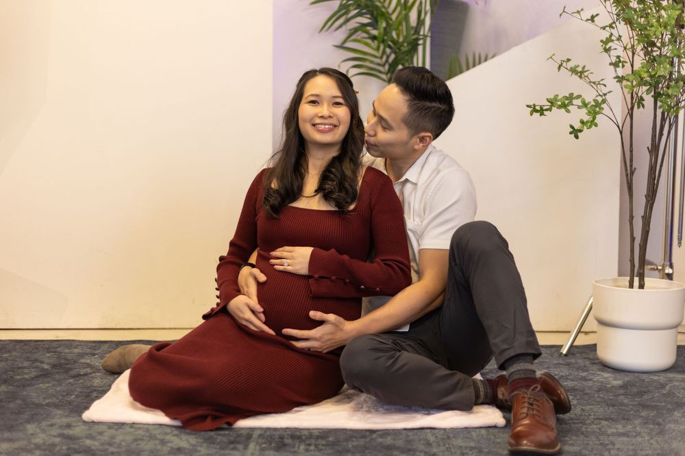 A pregnant woman in a red dress sits with her partner during a maternity photo shoot indoors.