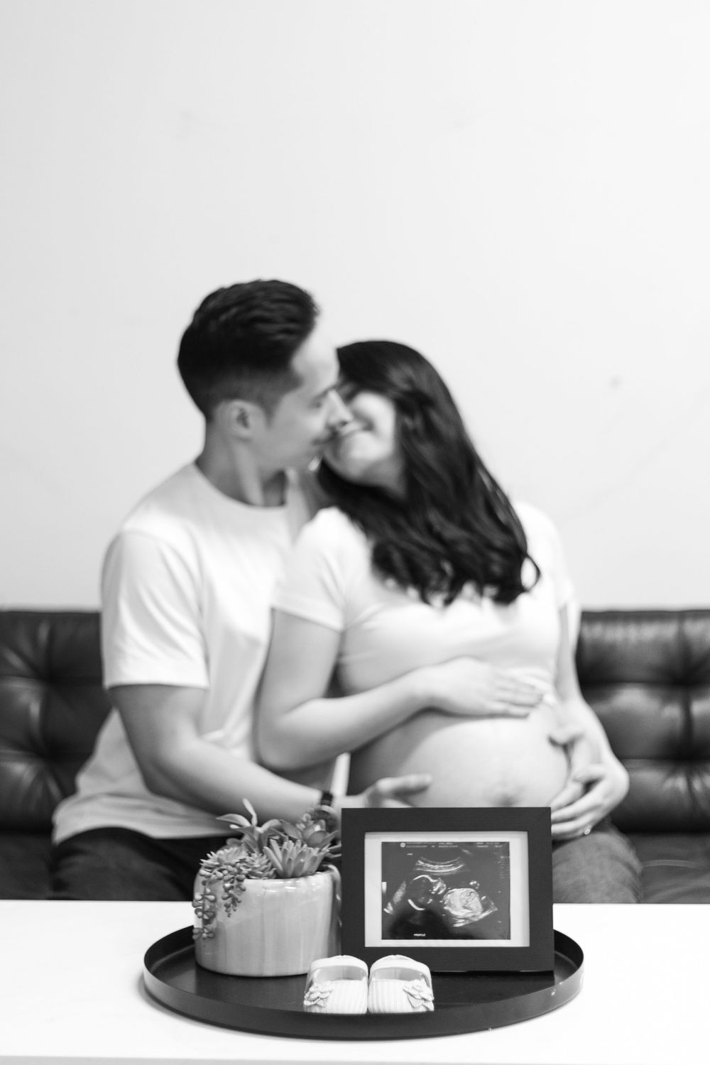 Black and white photo of pregnant couple kissing on sofa with ultrasound photo and baby shoes on tray in foreground.