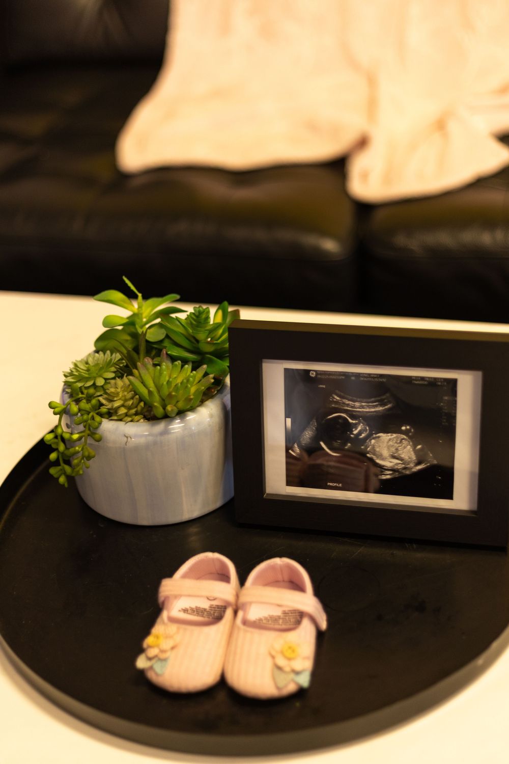Pink baby shoes and ultrasound photo in frame on tray with succulent plant, announcing a baby girl.
