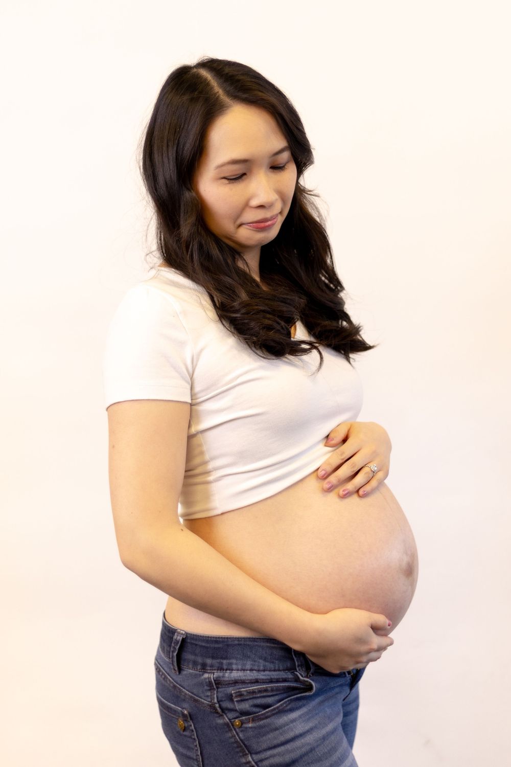 Pregnant Asian woman in white crop top and jeans cradling her baby bump against a white background.