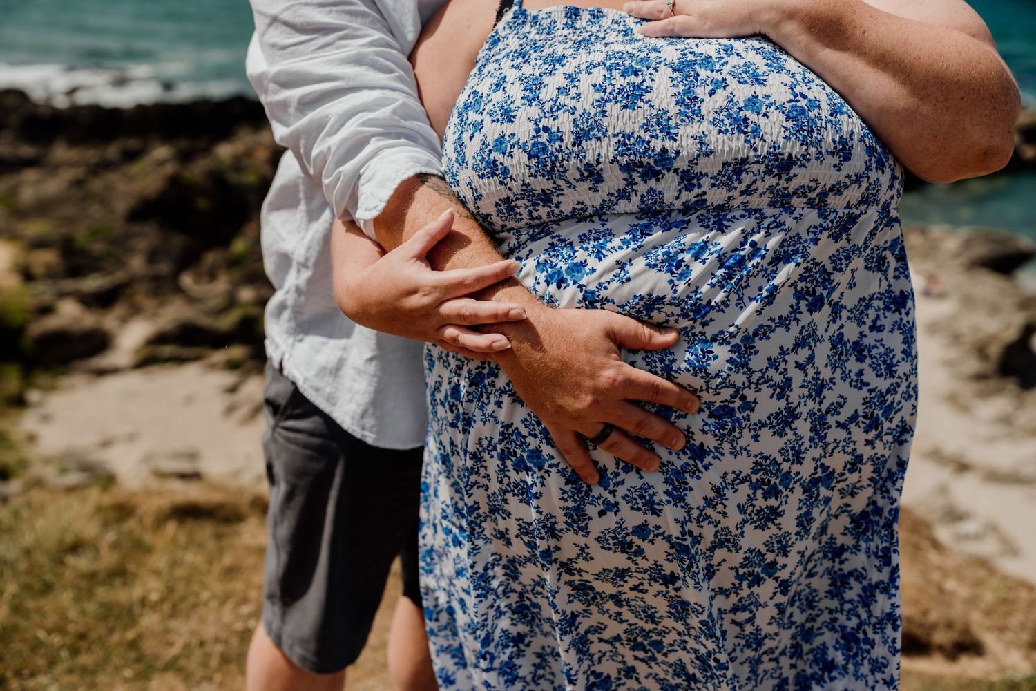 Couple embracing outdoors, hands cradling pregnant belly in blue floral dress near rocky ocean shore.