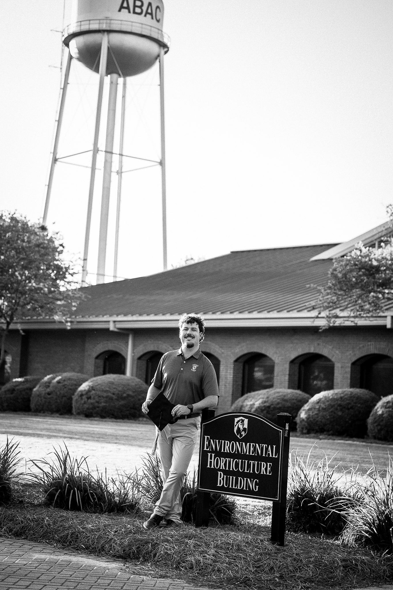 Man standing beside Environmental Horticulture Building sign with ABAC water tower in background, black and white photo.