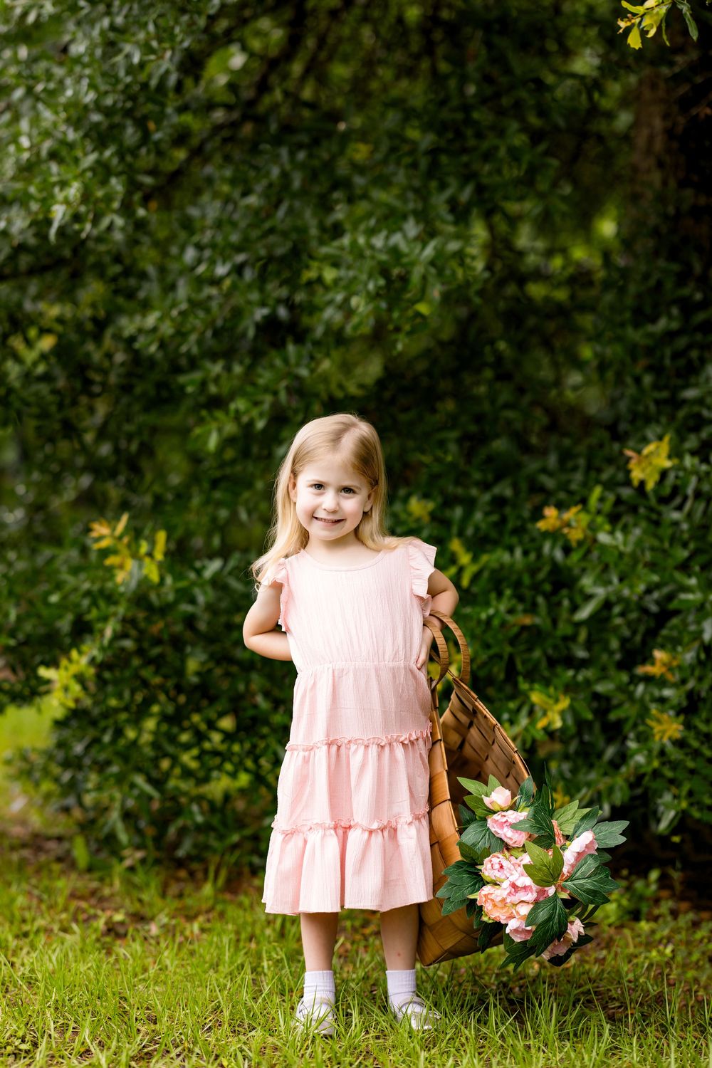 Young girl in pink dress holding floral basket, smiling outdoors among lush green trees.