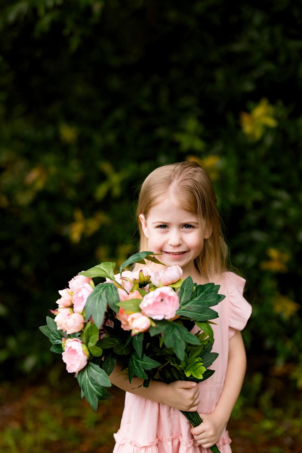 Young girl in pink dress holding a bouquet of pink peonies, smiling outdoors with green foliage background.