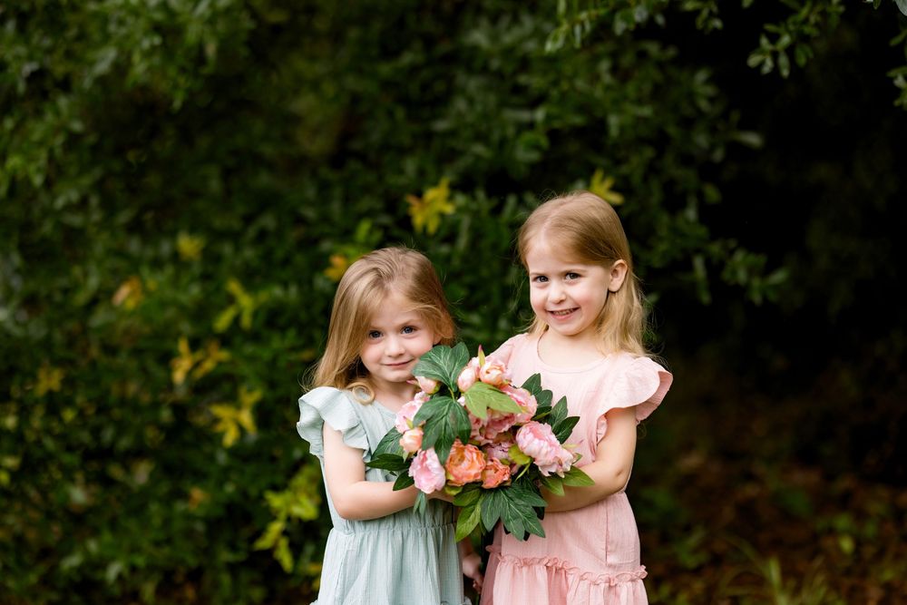 Two young girls in pastel dresses holding a pink floral bouquet outdoors among lush green foliage.