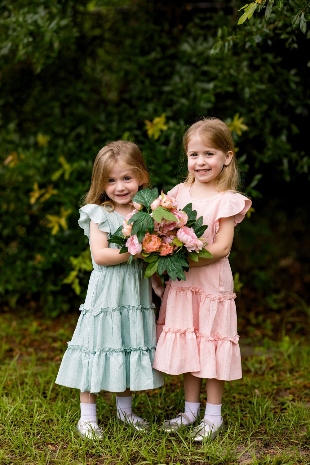 Two young girls in ruffled dresses holding a pink flower bouquet while smiling outdoors in a garden.