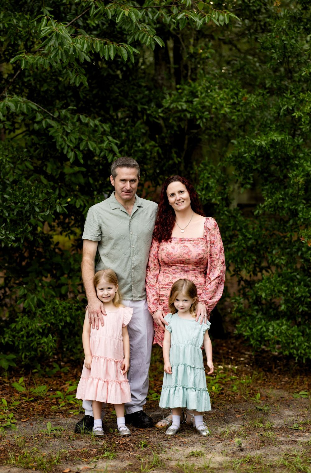 Family of four posing outdoors among lush green trees, parents with two young girls in pastel dresses.