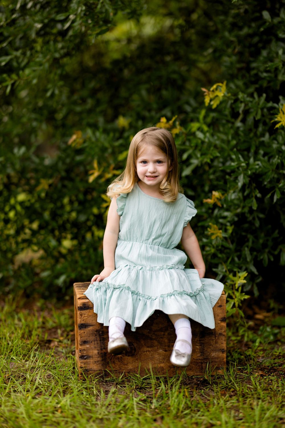 Young girl in mint green dress sitting on rustic wooden crate outdoors surrounded by lush green foliage.