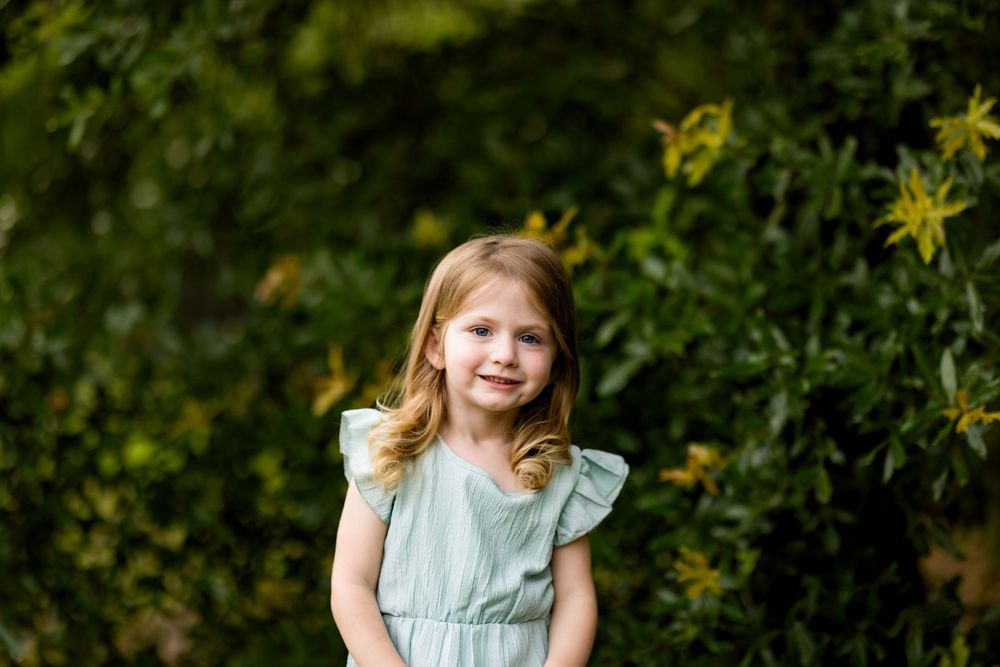A smiling young girl in a mint green dress poses outdoors among lush green bushes with yellow flowers.