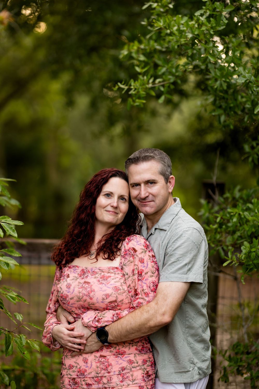 A couple embraces outdoors, woman in pink floral dress with red hair, man in gray shirt, surrounded by lush green trees.