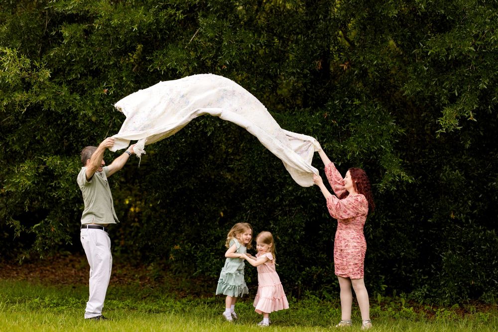 Family of four playing with white fabric outdoors, two parents lifting sheet over two young girls on green grass.