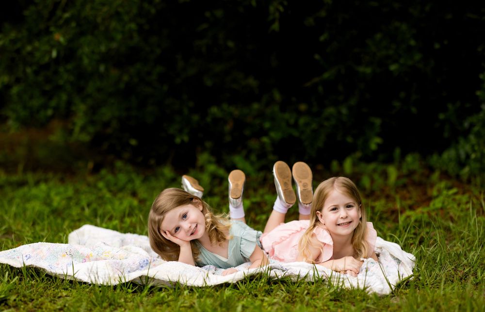 Two young girls lying on a blanket in green grass, smiling and posing with feet up in a sunny outdoor setting.