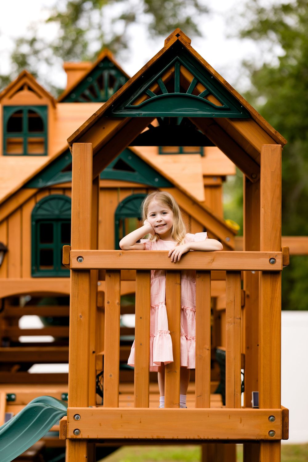 Young girl in pink dress smiling on wooden playground playset with green accents outdoors.