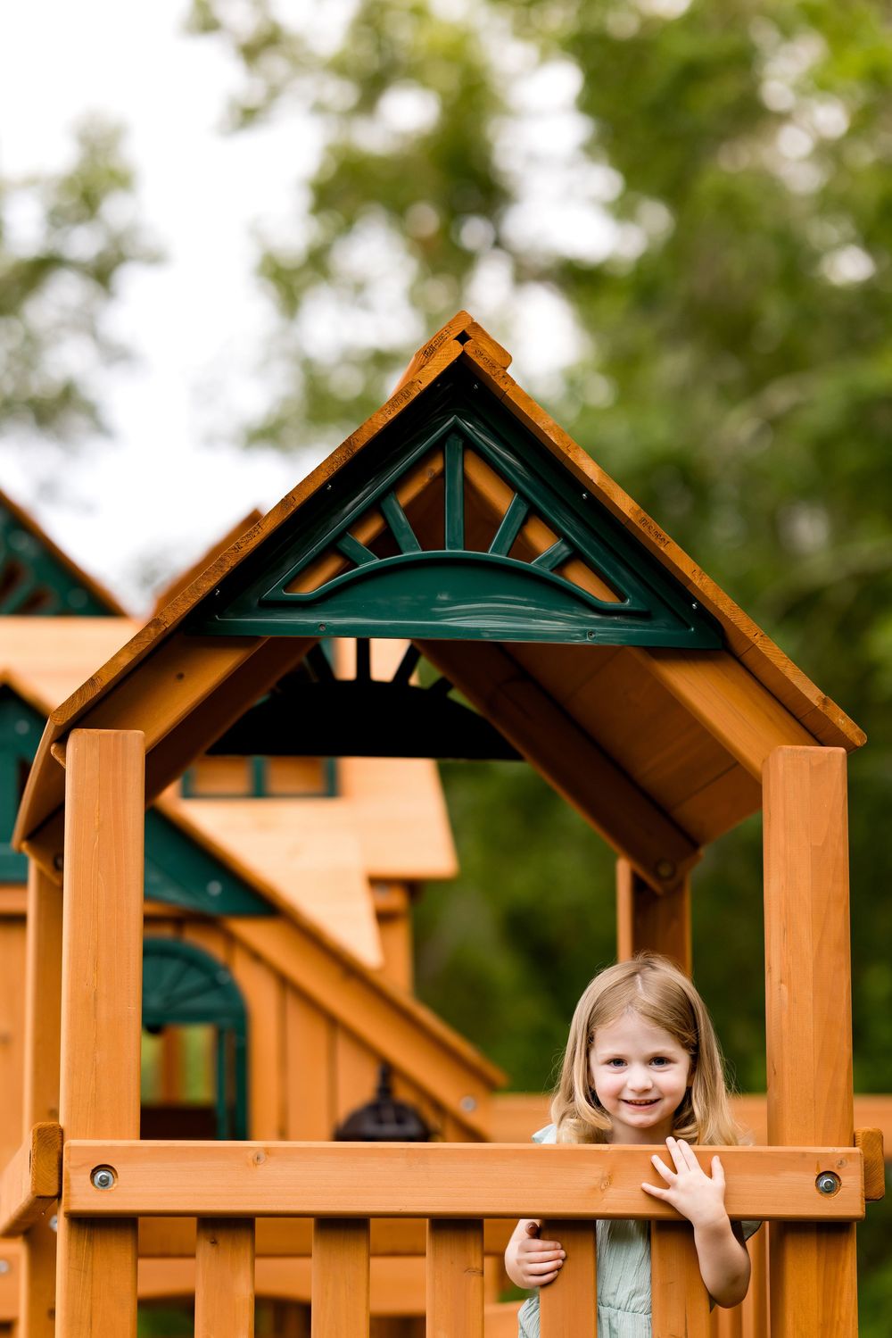 A smiling young girl waves from a wooden backyard playset with a decorative green and wood roof on a sunny day.
