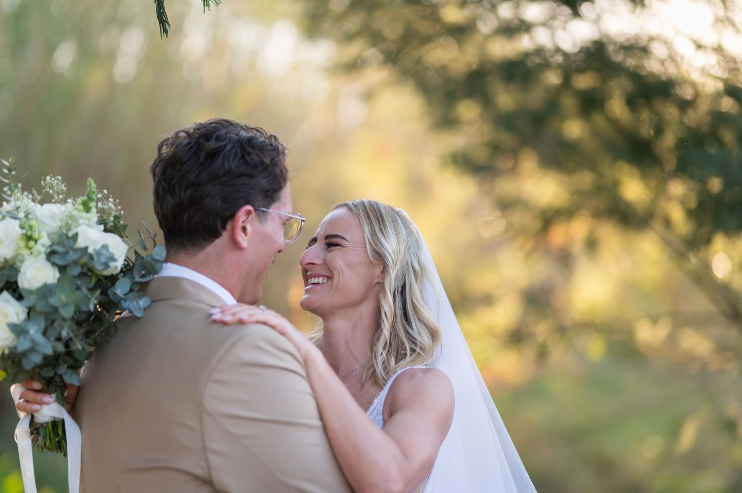 Bride and groom share a joyful moment outdoors, surrounded by lush greenery and golden sunlight on their wedding day.