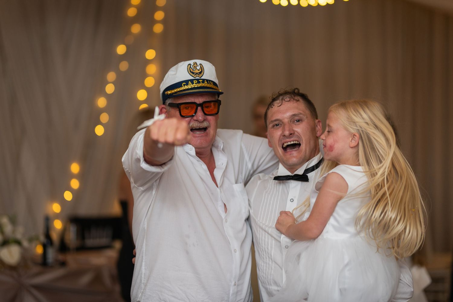 Man in suit hugging two young girls in white dresses at a wedding reception with fairy lights.