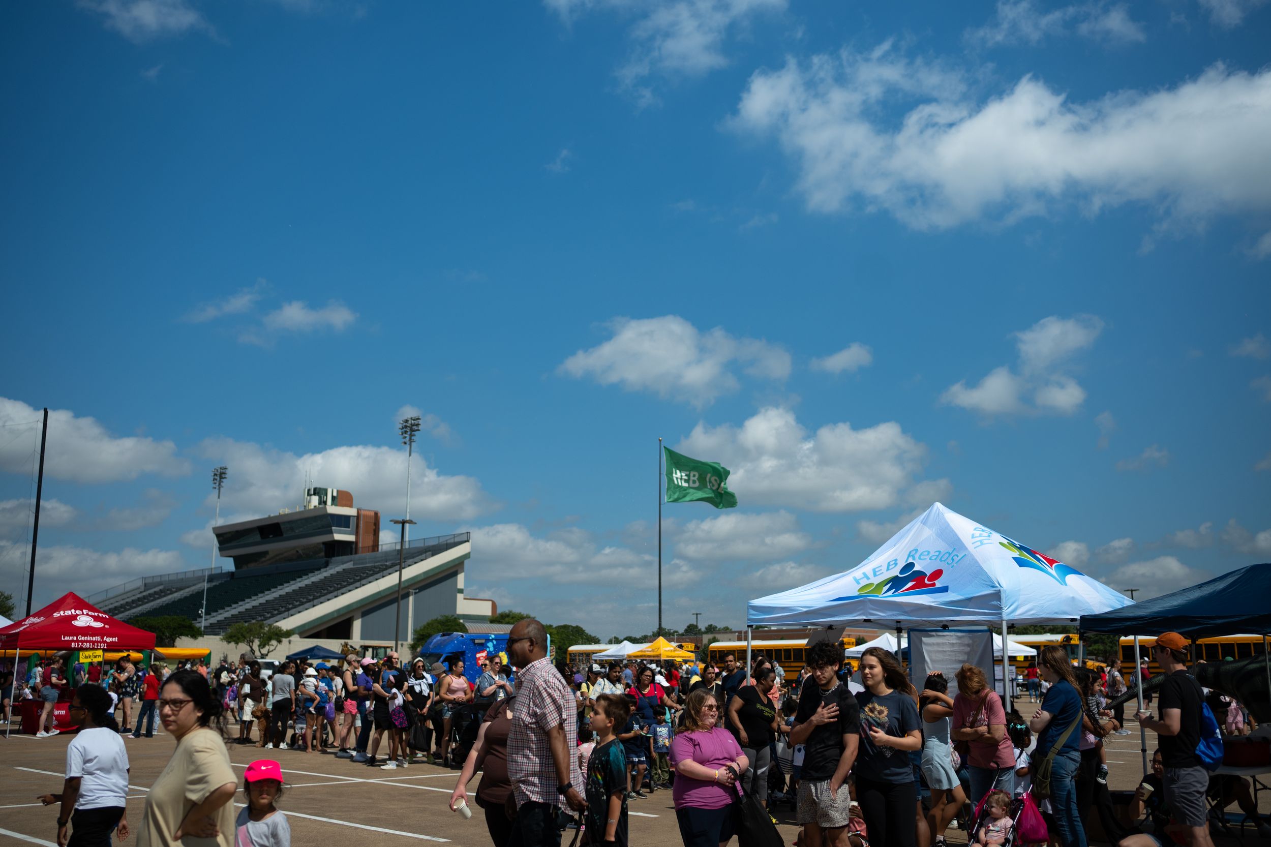 Crowded outdoor festival with colorful tents, green flag, and blue sky with clouds in background.