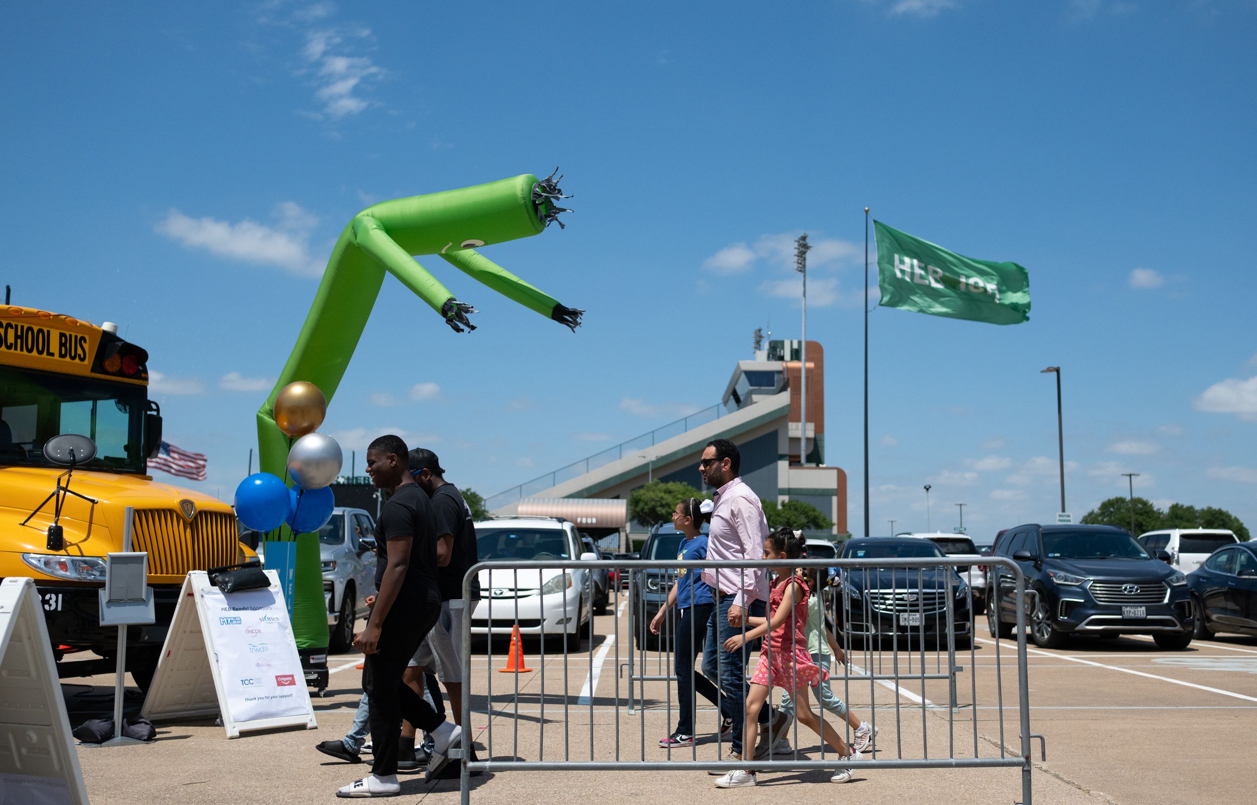 Outdoor event with green inflatable dancer, school bus, and flag in sunny parking lot.