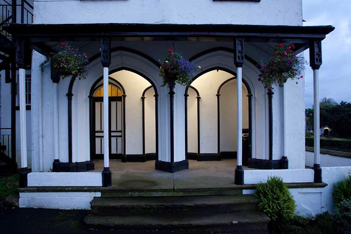 Elegant white building entrance with black-trimmed arches, illuminated columns, and hanging flower baskets at dusk.