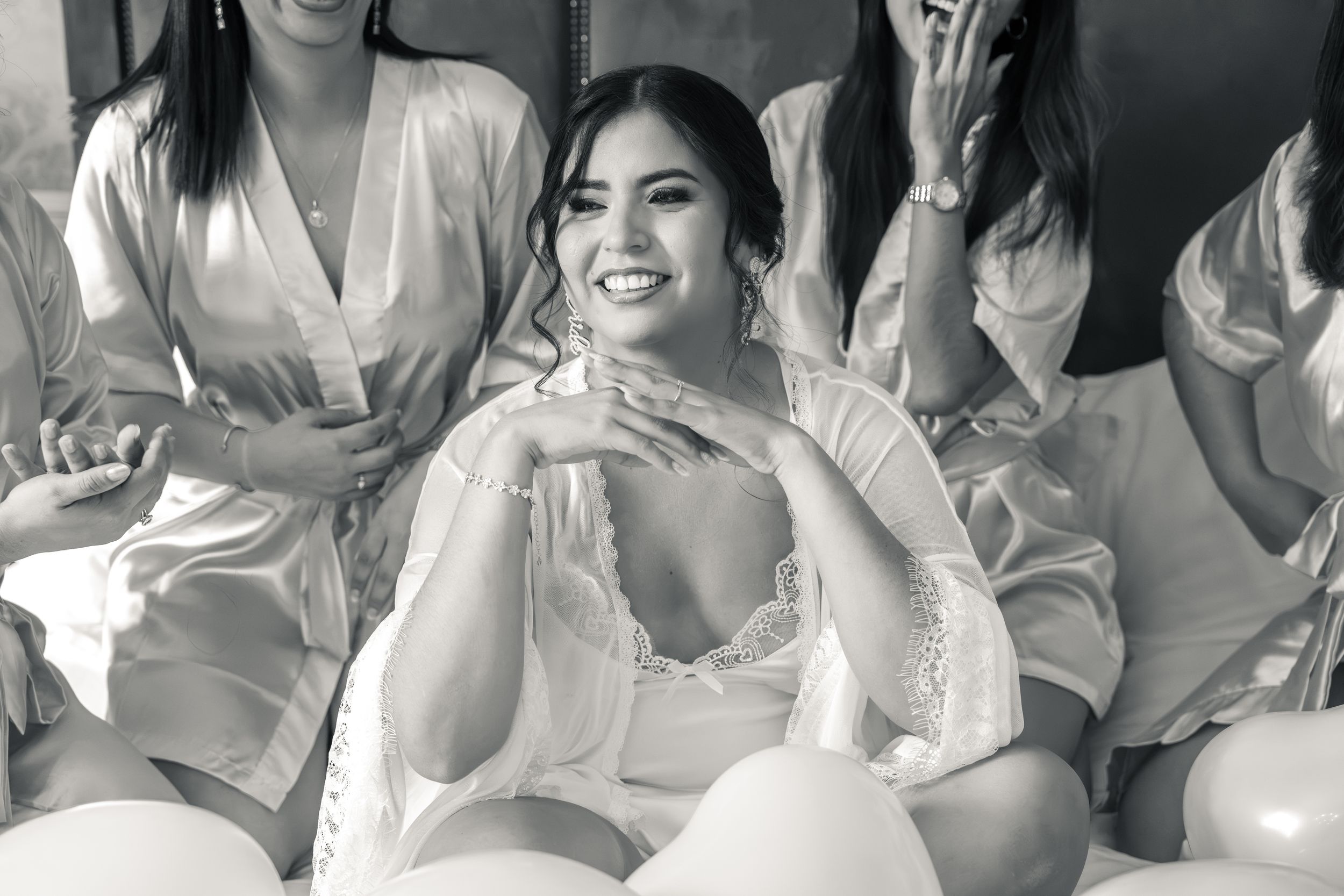 Black and white photo of a smiling bride in lace robe surrounded by bridesmaids in satin robes with balloons.