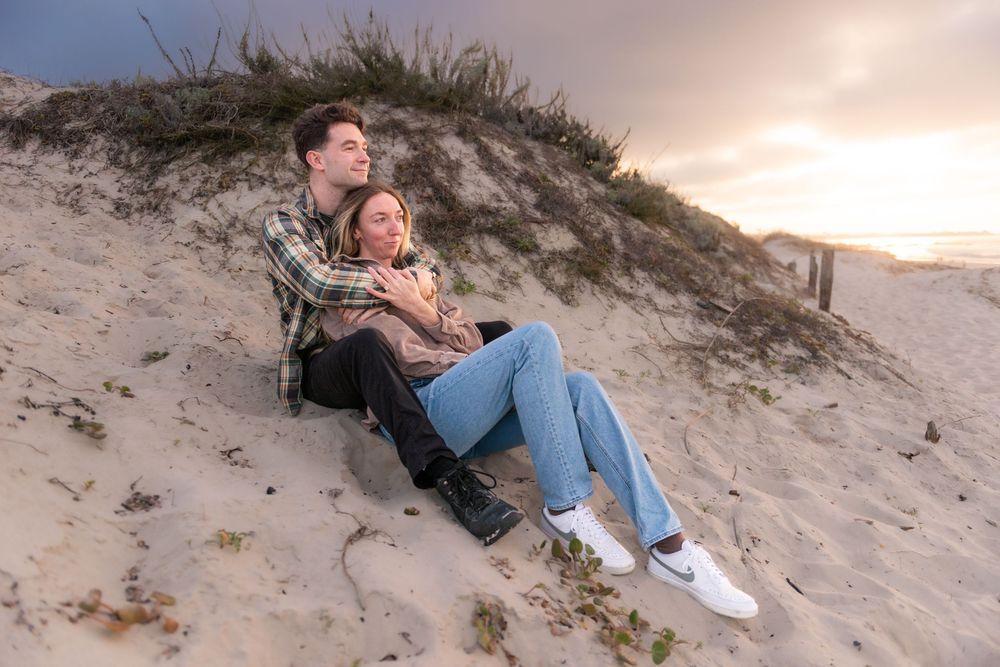 Couple sitting together on sandy beach dunes at sunset, wrapped in a blanket, enjoying a romantic coastal view.