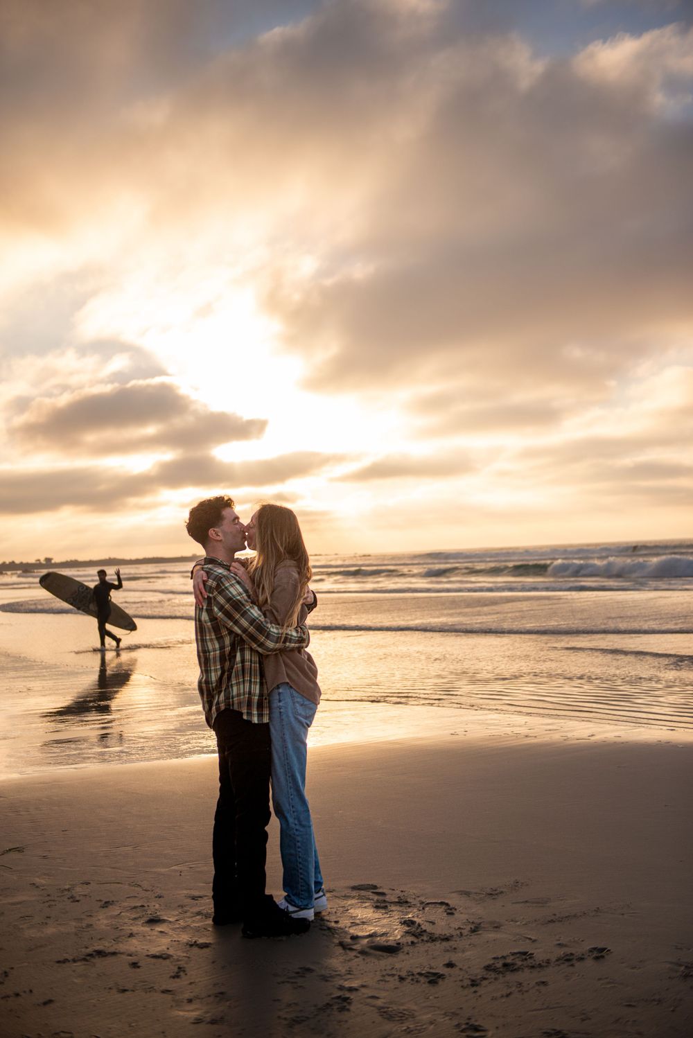 Couple kissing on a beach at golden sunset with a surfer walking in the background near ocean waves.