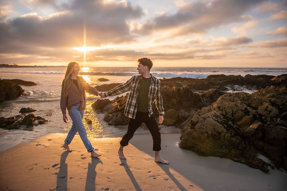 Couple holding hands walking on a sandy beach at golden sunset with rocky coastline and dramatic cloudy sky.