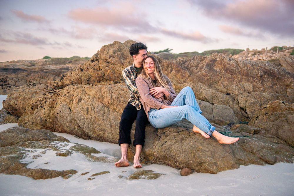 Couple sitting together on rocky beach at sunset, woman in jeans leaning back against man, romantic coastal scenery.