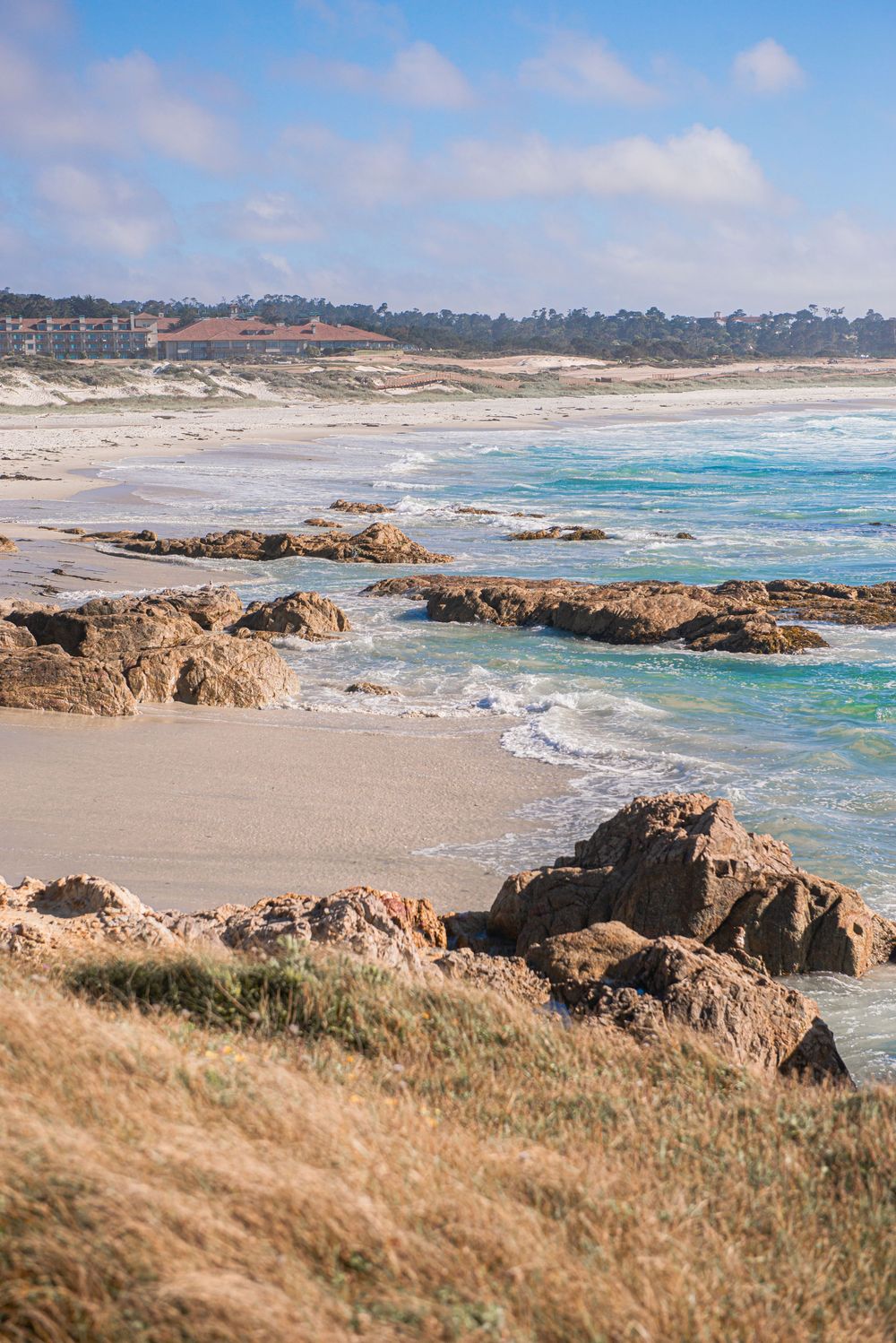 Rocky shoreline with turquoise waves, sandy beach, and coastal town in background under partly cloudy sky.